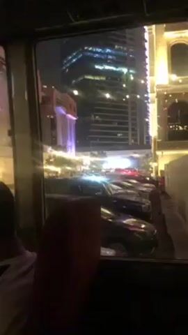 From inside a vehicle, a view is visible through the window. The view shows parked cars lining a street, buildings illuminated with lights, and a tall skyscraper in the background with blue and white accents. A person is sitting in the vehicle. Ambient conversation can be heard.