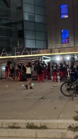 A nighttime scene shows a crowd gathered outside a modern building with large glass windows. The building has the word "LAFABRIKA" visible on its facade. A group of people, some wearing red pants and black shirts, are congregated near the entrance. One person is playing a drum. Other individuals are standing around, some walking. Trash and debris are scattered on the ground. In the background, trees and streetlights are visible. The lighting suggests it is dark outside, and artificial lights from the building and streetlights illuminate the scene. The overall atmosphere appears to be a public gathering or event.