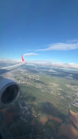 The video is a first-person view from an airplane window, looking out over a landscape below. The airplane's wing and engine are visible in the foreground, with the wing extending to the left of the frame and the engine to the lower left. The wing has a red logo on its tip. Below the airplane, a sprawling green forest is visible, dotted with buildings and roads, indicating a town or city. The sky above is a clear blue with scattered white clouds. The lighting suggests it is daytime, likely morning or afternoon, given the bright sunlight and shadows cast on the landscape. There are no people visible in the video. The overall scene depicts an aerial view of a landscape from a moving aircraft.