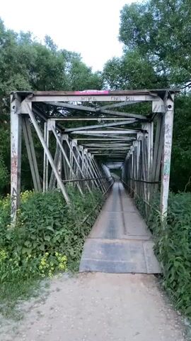 A drone flies over a narrow metal truss bridge. The bridge has a metal walkway with a railing on either side and is surrounded by lush green vegetation. The sky is overcast.