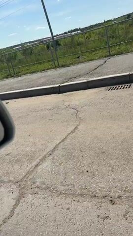 The video shows a view from inside a vehicle, looking out the passenger side. The camera pans slightly to the right. In the foreground, there is a rough, cracked asphalt surface with some debris. A concrete curb separates the asphalt from a patch of green grass and weeds. Beyond the grass, there is a paved path or road. Further in the background, a metal fence runs horizontally across the frame, with lush green vegetation behind it. In the distance, there are some buildings or structures and a clear blue sky with a few scattered clouds. The lighting suggests it is daytime.