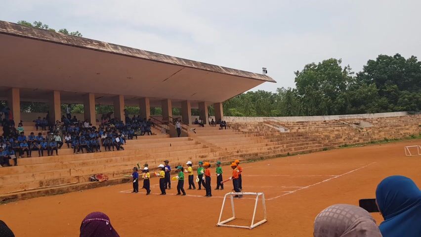 This media file captures an outdoor sporting event, likely a children's cricket match, taking place in Kalapet, India. The scene unfolds on a reddish-brown dirt field, characteristic of many cricket grounds. A tiered grandstand, constructed of concrete, rises behind the field, filled with spectators, many of whom are young people wearing blue uniforms, suggesting a school or youth event. The grandstand has a large, overhanging roof providing shade.

On the field, a group of children, dressed in brightly colored uniforms (yellow, green, orange, and blue), are participating in what appears to be a playful or instructional cricket game. Some are holding cricket bats and balls. The children are positioned in a line, facing towards the spectator stands, and seem to be engaged in a demonstration or practice.

In the background, lush green trees are visible, indicating a warm climate. The sky is overcast, suggesting a potentially humid day, but not a harsh sun. The overall atmosphere seems to be one of community gathering and youthful sporting activity. The presence of adults in the spectator stands and observing from the sidelines suggests organized supervision. The image is taken from a slightly elevated perspective, looking down onto the field and the participants. No visible text is present in the media.