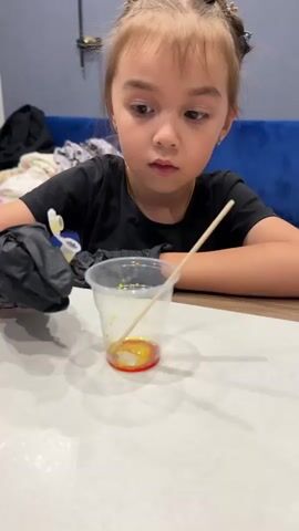 A young girl, likely under ten years old, sits at a table wearing a black t-shirt and black gloves. She is focused on a clear plastic cup containing a red and yellow liquid. A wooden stirring stick is in the cup. The girl is holding a small yellow dropper bottle and is in the process of adding liquid from the dropper into the cup. Her hair is styled in two buns with decorative clips. The background is slightly blurred, showing a blue couch and some fabric. The scene appears to be indoors, with diffused lighting suggesting it could be daytime. The girl's expression is one of concentration. There is no visible text in the media. The location context is Murom, Russia.