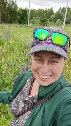 A woman is filmed outdoors in a field of purple flowers. She is wearing a green jacket, a patterned shirt, a baseball cap, and sunglasses. She smiles at the camera, then picks a flower and smells it. The background shows a grassy field and trees.