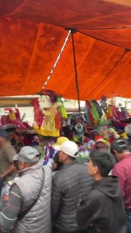A crowded outdoor market is depicted under a large orange tarp. Several people are visible, some wearing hats and casual clothing. In the center, a group of individuals appear to be performing, with one person holding up cymbals. Colorful feathered headdresses and costumes are prominently displayed. The atmosphere seems festive, with a band playing instruments, including what appears to be a brass instrument.
