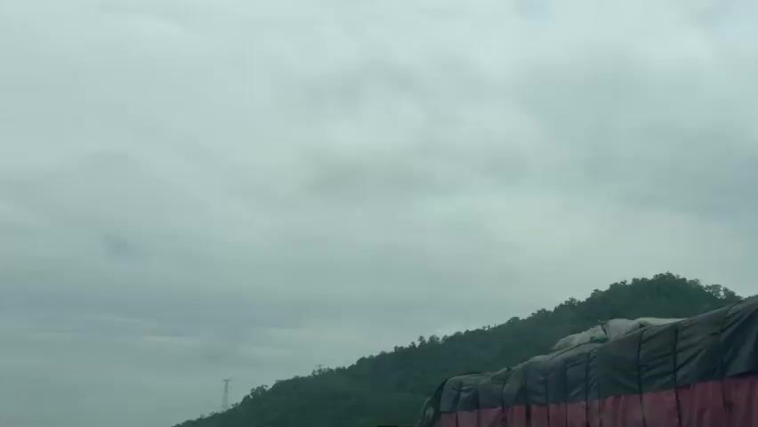 The video shows a view from a vehicle traveling on a two-lane road. The road is asphalt and appears to be in fair condition. On the right side of the road, there are several large trucks parked, many of them covered with tarpaulins. A person in a red dress is standing next to one of the trucks. On the left side of the road, there are some small buildings, piles of sand or gravel, and some utility poles. The landscape behind the road consists of a lush green, tree-covered mountain under a cloudy sky. The vehicle is following a black pickup truck with a white cap, and other vehicles are visible further down the road. The overall impression is of a busy highway with commercial traffic, possibly in a rural or semi-urban area.