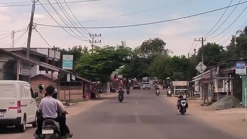 The video shows a straight, paved road in a rural or semi-rural area, with a moderate amount of traffic consisting of motorcycles and a white SUV. The road is lined on both sides with small, simple structures that appear to be shops or stalls, many of which have awnings or tarps. Power lines are visible overhead, crisscrossing the road. Trees are present, adding a touch of greenery to the scene. People are visible riding motorcycles and standing near the shops. The sky is slightly overcast.