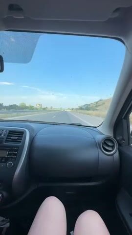 This is a first-person point-of-view video taken from inside a car driving on a highway. The view is directed forward through the windshield. The dashboard of the car is visible in the foreground, showing a black plastic surface with air vents, a radio, and a control knob. Below the dashboard, the legs of a person are visible, wearing what appears to be shorts. The highway stretches out ahead, with a clear blue sky and scattered clouds above. The sides of the highway are lined with dry vegetation and hills. There are some faint smudges or dirt marks on the windshield. Other vehicles are visible in the distance on the highway. The lighting suggests it is daytime, likely in the afternoon or early evening, with the sun casting a bright light.