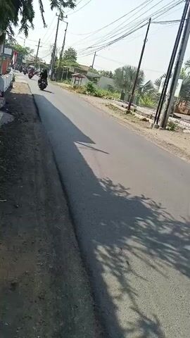 The video shows a road with a few motorcycles on it. The road is paved and has a dirt shoulder on the left. There are trees and buildings on the sides of the road. The sun is shining and casting shadows on the road. The video appears to have been shot from a moving vehicle.