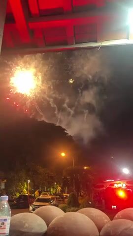The video captures a nighttime scene of fireworks being set off. In the foreground, out of focus, are round, dome-shaped objects, possibly food items or decorative elements. A plastic water bottle is visible on the left. The middle ground shows a street with cars parked and streetlights illuminating the area, with trees lining the street. The background is dominated by a dark sky filled with bursts of bright red and yellow fireworks. Red wooden beams are visible at the top of the frame, suggesting the viewpoint is from under a structure, possibly a roof or awning. The fireworks display appears to be large-scale, with multiple explosions in the sky, creating clouds of smoke and light.