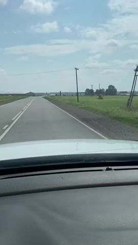 The video shows a view from inside a vehicle, looking out the windshield at a straight, paved road with double white lines. The road is bordered by green grassy fields on the right, with scattered trees and power lines in the distance. The sky is blue with scattered white clouds. The dashboard of the vehicle is visible in the foreground. The vehicle appears to be moving forward at a moderate speed.