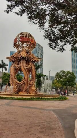 The video shows a golden dragon fountain in a plaza. The fountain features multiple dragons intertwined, supporting a wireframe globe with "SUNWORLD HALONG" written on it. Water cascades down from the globe and the dragons into a circular pool. The plaza is paved with interlocking bricks, and a few people are walking around in the background. Behind the fountain, there are buildings, trees, and a cloudy sky.