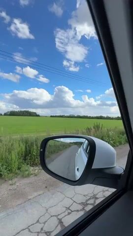 The video shows a view from inside a car, looking out the passenger side window and into the side mirror. The car is on a dirt road next to a green field. The sky is blue with fluffy white clouds, and power lines stretch across the sky. The side mirror reflects the road behind the car, showing the white car continuing down the dirt path. The overall impression is one of a scenic drive through the countryside on a bright, sunny day.