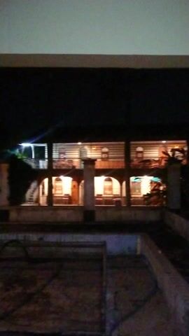 The video, filmed at night, shows a dark environment, likely looking out from a building or apartment. The camera slowly pans downwards, revealing another building or apartment complex across a dark void. Several windows and balconies of this opposing structure are visible, with lights illuminated inside, indicating human activity. A light-colored wall or pillar is prominent on the left side of the frame throughout the video, and another structural element is visible on the right. No people or text are discernible.