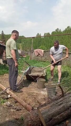 In an outdoor daytime scene, two men are engaged in a construction-related activity. One man, wearing a light green t-shirt and dark gray pants, stands observing. The other man, clad in a light gray t-shirt and black shorts, is mixing cement in a bucket using a shovel. A wheelbarrow filled with cement sits nearby. The environment includes a wooden fence, trees in the background, and various construction materials such as logs and pipes scattered around. A rug hangs on the fence. The sky is partly cloudy.