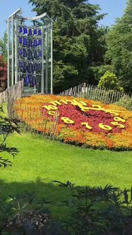 A flower clock in a park has the numbers for 7 o'clock visible. There are blue bells on display in a glass structure.