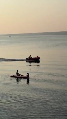 The video shows a wide body of water with a hazy sky. In the foreground, two people are on a paddleboard. One person is sitting on the paddleboard, and the other is kneeling behind them. In the background, a small red inflatable boat with an outboard motor is moving across the water, leaving a wake. There are two people in the boat. In the far distance, a large cargo ship or barge is visible on the horizon. The water is calm with gentle ripples. The overall lighting suggests it is either sunrise or sunset.