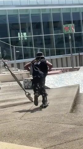 A person is seen from the back, riding a black scooter across a concrete plaza area under bright daylight. The rider is wearing a dark, sleeveless top, patterned shorts, a dark hat, and dark shoes. They approach and ride up a concrete structure that features a white tiled surface, resembling a ramp or bank in a skate park. In the background, there is a large modern building with a facade made predominantly of large glass windows. To the left, a large metal beam or support structure is visible. A metal pole with a small green square sign is also in the background. The scene depicts an outdoor urban environment, possibly a skate park or public plaza, in Bron, France.