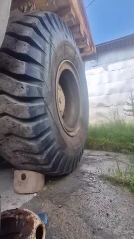 The video shows a close-up, ground-level view of a very large tire on a heavy-duty vehicle. The tire is black with a thick tread pattern, and it is resting on a light-colored block. The vehicle appears to be a mining truck or a similar piece of industrial equipment. The background shows a dirt or gravel surface, some green vegetation, and a building or structure with a white corrugated metal exterior. The lighting suggests it is daytime. The camera angle is low, emphasizing the immense size of the tire. There are no people visible in the video. The overall scene depicts a parked piece of heavy machinery.