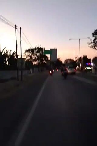 This is a video taken from the perspective of someone on a motorcycle, traveling down a two-lane road at dusk. The sky is a soft gradient of purple and orange. Several other motorcycles and a car are visible on the road ahead, traveling in the same direction. Trees and buildings line the side of the road, and a large green billboard is visible above the trees. Streetlights are also present along the road. The overall atmosphere is peaceful, with the soft lighting of dusk creating a serene scene.