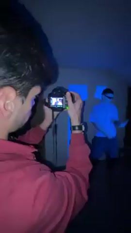 A person in a red shirt is holding a camera and filming another person in a light-colored shirt and dark shorts. The person being filmed is wearing a visor over their eyes and moving their arms. There is a square pattern of light projected on the wall behind them. The lighting in the room is dark.