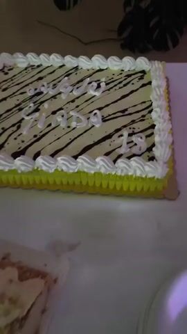 A rectangular cake with white icing and yellow trim is decorated with diagonal dark lines and white lettering. The cake has "Feliz Cumple" written in white script on the top left, and "Giada 15" in white script on the bottom right. The cake is on a white tablecloth, and there are some utensils on the table. There are other desserts in the background, including a round cake with meringue peaks and a long rectangular pastry with cream and fruit. The environment appears to be a party or celebration.