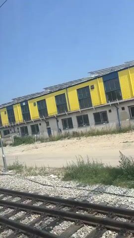 The video shows a view from a train moving along a railway track. The camera is angled slightly upwards, capturing a bright blue sky with a few faint clouds. Adjacent to the tracks, there is a dirt path and a grassy area with scattered weeds and bushes. Beyond the path, a row of modern, industrial-style buildings is visible. The buildings are mostly painted in bright yellow and grey or blue and yellow, with visible solar panels on their roofs. Some of the buildings have large windows, and one appears to be a warehouse or factory. A tall, grey lamppost stands in the foreground, and further along, a train signal pole with black and yellow markings is present. There are no people visible in the video. The overall impression is that of a semi-industrial area on a sunny day, seen from the perspective of a passing train.