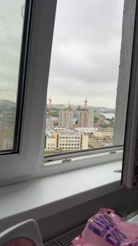 The video pans across a city skyline from inside a building. The foreground shows a windowsill and a portion of a window frame. Beyond the window, the view includes construction sites with cranes, residential buildings, industrial buildings, a body of water, and a large cable-stayed bridge in the distance. The sky is overcast. The overall impression is of an urban environment undergoing development, with a mix of older and newer structures.
