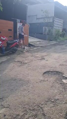 Here is a concise description of the video:

The video shows an outdoor scene with a young man and a toddler. The man is standing, looking at his phone, while the toddler walks around him on a dirt road. A red scooter is parked nearby. The area appears to be a residential neighborhood, with buildings and greenery in the background. The road is uneven and has some puddles. The lighting suggests it is daytime.