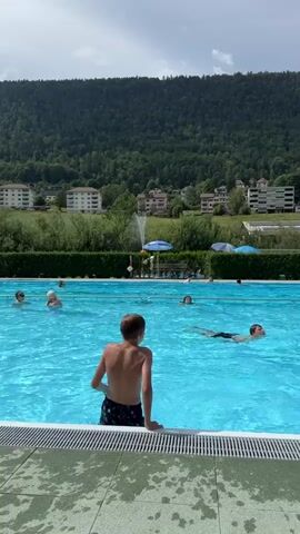 A group of people are swimming in an outdoor swimming pool. The pool is blue and surrounded by a tiled deck. In the background, there are trees, buildings, and a mountain. The sky is cloudy. The video starts with a boy sitting on the edge of the pool, facing away from the camera. He then enters the water and swims. Other people are also swimming and playing in the pool. The video ends with the boy swimming in the pool.