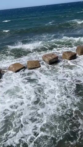 The video shows a choppy, dark blue ocean with white waves crashing against square, concrete blocks. The blocks are spaced evenly apart and appear to be forming a barrier or part of a structure. The water is foamy and turbulent as it churns around the blocks. The sky is a clear, bright blue.