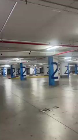 The video shows the interior of a large, mostly empty parking garage. Rows of white columns painted with a large blue number "2" support the ceiling. Red pipes run along the ceiling, and there are overhead lights. The floor is grey concrete. The camera slowly moves forward between the rows of columns. The video is shot with a slight upward tilt.  The scene is lit with bright, artificial light.