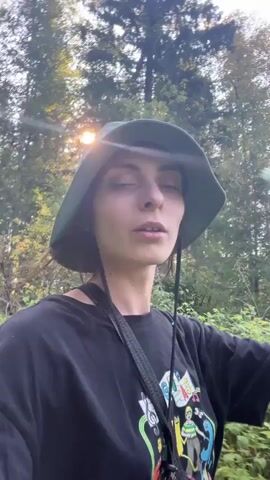 A person wearing a green bucket hat and a black t-shirt with a colorful graphic print is walking in a wooded area. The person is holding a phone to record a video of themselves. Sunlight filters through the trees, creating a dappled effect on the person's face and the surrounding foliage. The person's expression is neutral, and they appear to be looking directly at the camera. The video is shaky, indicating that the person is in motion. The background is a blur of green leaves and trees, suggesting a natural environment.
