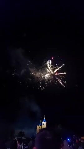 The video shows a nighttime fireworks display. In the foreground, silhouetted heads of a crowd are visible. In the middle ground, a building resembling a castle or church with turrets and spires is illuminated by the fireworks. The building has a pale yellow facade and blue spires. In the background, fireworks are exploding in the dark sky, creating bursts of light and smoke. Some fireworks are white and yellow, while others are pink and red. The fireworks are positioned above and behind the building.