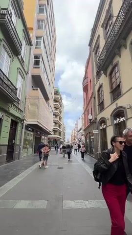 The video shows a pedestrian walkway lined with shops and buildings in what appears to be a city. The buildings vary in height and are painted in a variety of colors including green, pink, yellow, and cream. Many people are walking along the walkway. Some appear to be couples or in small groups. The sky is visible above the buildings and is partly cloudy. The walkway is paved with grey blocks.