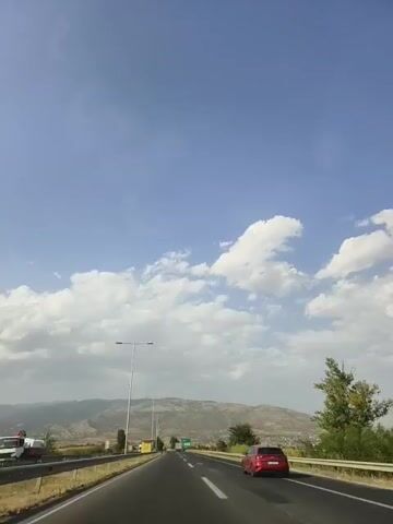 The video shows a view from inside a vehicle traveling on a highway. The sky is blue with scattered white clouds. There are mountains in the distance. A red car is visible on the right side of the highway, and a truck is visible on the left side. There are streetlights and signs along the road.