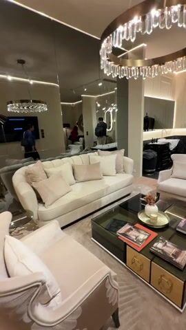 The video shows a room with a combined living room and bathroom display, likely in a showroom or store. In the foreground is a seating area with a cream-colored sofa, armchair, and a dark, multi-tiered coffee table. Above is a circular chandelier with crystal-like pendants. Behind the seating area, the space transitions into a bathroom setup, featuring a black vanity unit with a sink, a black-framed bathtub with white interior, and a toilet. The bathroom area is lined with black marble-like material and has a large mirror. Overhead, a different, more modern chandelier hangs. Several people are seen walking through the space, seemingly as customers or visitors.