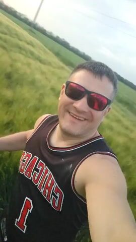 The video shows a man wearing a black Chicago Bulls jersey and sunglasses, standing in a field of tall green grass. He is smiling and looking at the camera. The background is blurry, but it appears to be a field and a road. The sky is white, suggesting a bright or overcast day.  The camera angle is slightly canted, as if the person is holding the camera at an angle to take a selfie video.
