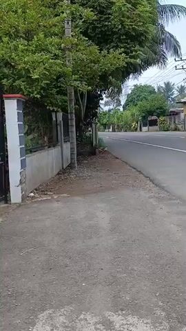 A motorcycle passes by on the right side of the road. A wall and trees are on the left side of the road. The road is paved and has white lines. There are houses on the right side of the road. The sky is cloudy.