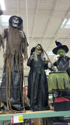 A video taken from a slightly low angle shows three large Halloween decorations on a shelf in what appears to be a retail store. The decoration on the far left is a tall, skeletal figure draped in a tattered brown robe with a black base. Its skull-like head emits a blue glow from its eye sockets. In the center is a witch decoration wearing a black dress and holding a staff, with a black crow perched on its shoulder. To the right is another witch decoration in a black dress and a pointed hat, stirring a large green cauldron. Below the shelf, price tags are partially visible, one showing $229.99. The background shows the ceiling of the store with exposed beams and lighting fixtures. The overall scene is a display of Halloween props.