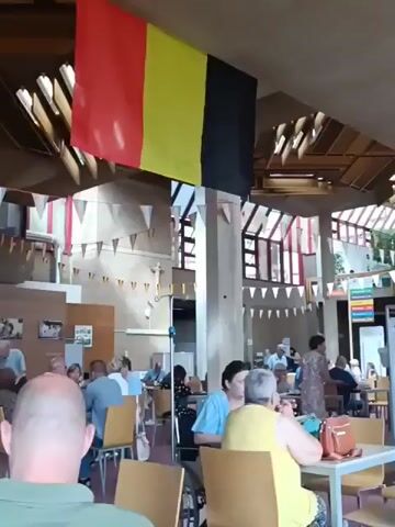 A Belgian flag hangs from the ceiling of a brightly lit room. Several people are seated at tables, some appearing to be elderly. A woman in a yellow shirt is seen from behind, facing two other people at her table. Another woman in a blue shirt is also at the table. A wheelchair is parked beside the woman in the blue shirt. In the background, more people are seated at tables, and there are decorations that appear to be small flags strung between poles. The room has high ceilings with exposed beams and large windows that allow natural light to enter. A service cart is visible on the right side of the frame.