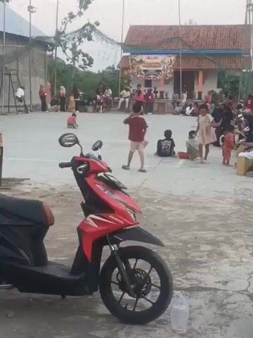 A red Honda scooter is parked in the foreground of the image. The background shows an outdoor court area where a group of people, including children and adults, are gathered. There is a building with a banner in the background and a fence with netting above it. Some children are playing or running on the court, while others are sitting or standing around. The overall scene appears to be a community event or gathering.