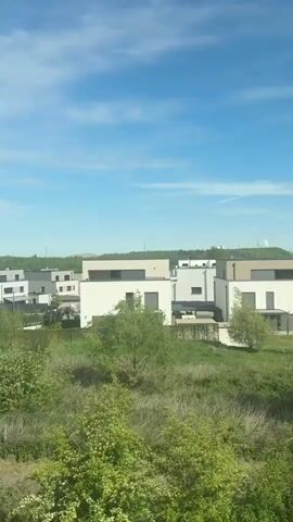 The video captures a panoramic view of a modern residential area on a clear, sunny day with a blue sky and scattered clouds. The camera pans from left to right, suggesting movement, possibly from a train or a high vantage point.

In the foreground, lush green foliage, including bushes and trees, lines the lower part of the frame. A paved path winds through this greenery. As the camera moves, a children's playground with climbing structures on a yellow ground cover becomes visible, followed by a fenced sports court, likely for tennis or basketball, surrounded by green fencing.

The midground is dominated by contemporary residential buildings, mostly two-story structures with white and grey facades, large windows, and flat or low-pitched roofs. These buildings are interspersed with more green spaces, including trees and lawns.

In the background, beyond the residential cluster, there are distant hills or industrial mounds, with some larger buildings or industrial structures faintly visible on the horizon. The overall impression is one of a well-planned, modern suburban or exurban development blending with natural green areas under a bright sky. There are no people visible in the scene.
