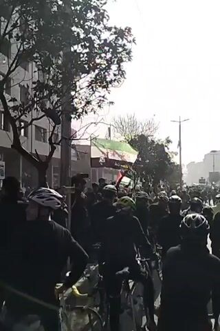 The video shows a large crowd of people, many wearing bicycle helmets, participating in what appears to be a procession or demonstration. Some individuals are holding flags. The scene is outdoors on a city street, with buildings visible in the background. The lighting is bright, with the sun visible in the sky, which could suggest a daytime setting. The overall atmosphere seems lively and potentially politically charged, given the presence of flags.