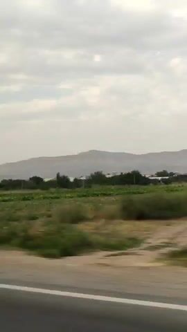 The video shows a view from a vehicle driving on a road. The landscape consists of agricultural fields, some with green crops and others with dry grass. There are trees and some buildings, including houses, scattered throughout the scene. In the background, there are mountains under a cloudy sky. The camera is panning, indicating movement.