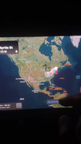A hand is seen interacting with a map displayed on a screen, likely a GPS navigation system. The map shows a route across the eastern United States, with various pins marking locations along the path. The route appears to stretch from the Midwest, through the Mid-Atlantic states, and down to the Southeast. The hand is in shadow, obscuring details of the person. The screen displays route information at the bottom, including distance and estimated travel time. The background is dark, suggesting the environment is dimly lit, possibly inside a vehicle at night.