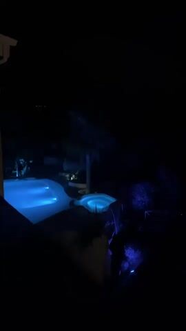 The video opens with a view of a dimly lit outdoor area at night. A swimming pool and a hot tub are illuminated with blue lights. As the camera pans, the view expands to show a city spread out in the distance, its lights twinkling against the dark landscape. The camera continues to pan and zoom out, revealing more of the city and the surrounding hills under the night sky.