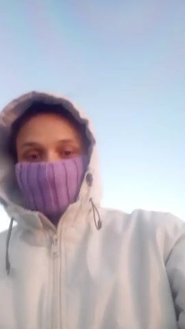 A person in a white hooded jacket and purple knit face covering stands in front of a bright sky, then turns to face the sunrise over a body of water. The lighting shifts dramatically. The sky is clear. The ground is rocky with pools of water.