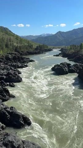 The video shows a river flowing through a mountainous landscape. The water is a light, murky color, and the river is turbulent, with white foam visible on its surface. Large, dark rocks line the riverbanks. Mountains covered with green vegetation rise in the background. Patches of snow can be seen on the distant mountain peaks. The sky is clear and blue with a few scattered white clouds. Laughter is heard in the background.
