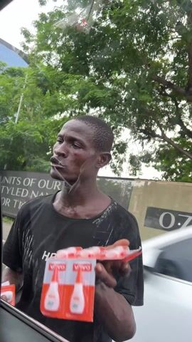 The video shows a man standing outside a car window, holding a display of VOVO-branded products. The man is wearing a black shirt with "HBA" written on it, and the shirt appears to be splattered with white paint. The man has a thin build and visible sweat on his neck and chest. He looks off to the side with a somewhat solemn expression. Through the car window, a white car is visible in the background, along with trees and a sign that reads "UNITS OF OFFICE". Another person walks in the background. The scene is outdoors, presumably during the day.